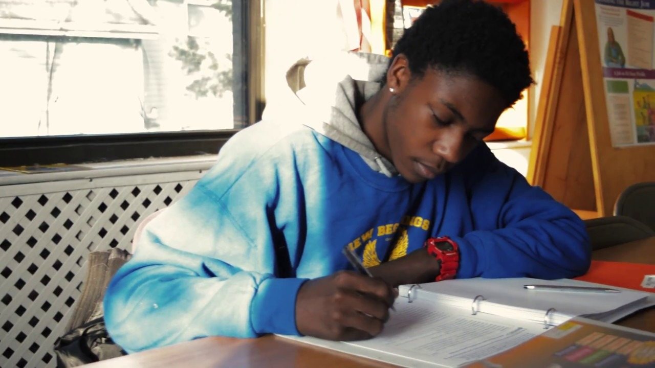 A student in a blue academy sweatshirt writing in a binder at a table by a window
