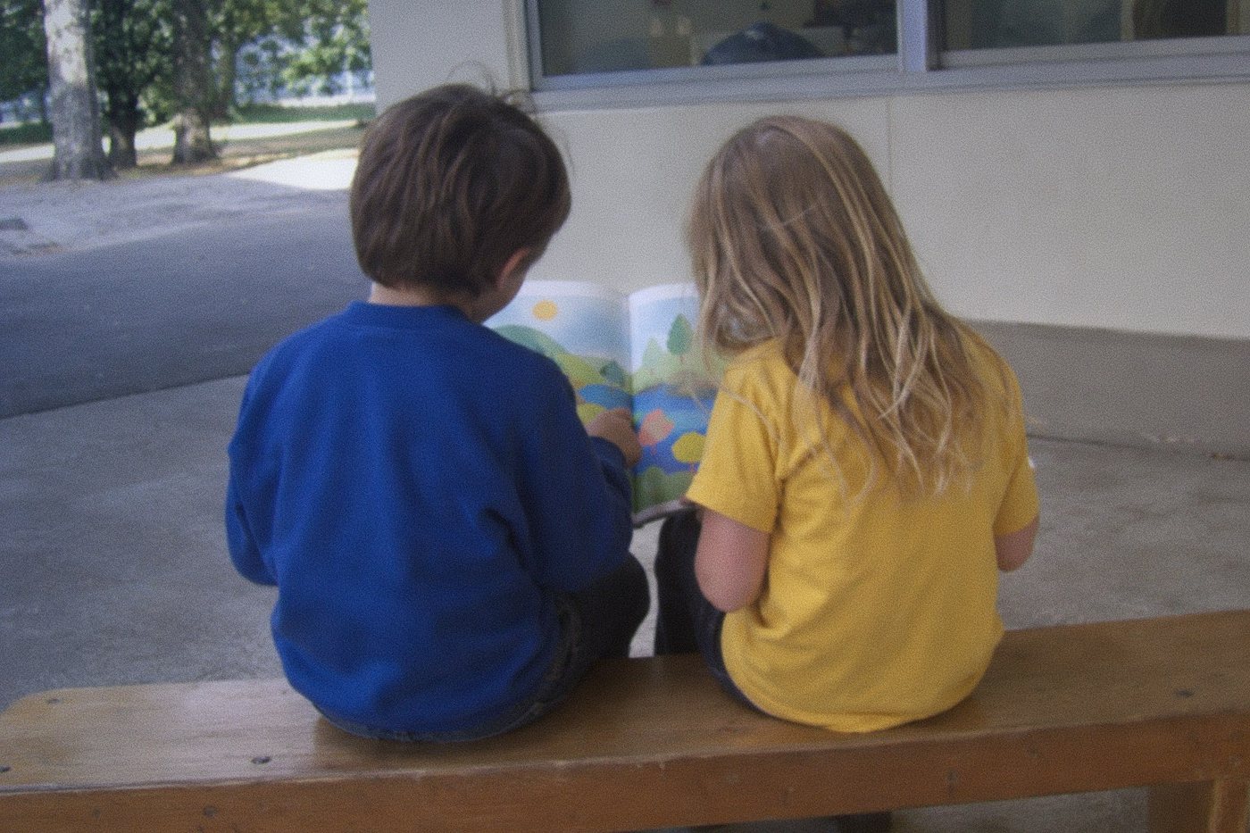 Two children, one in a blue sweatshirt and one in a yellow shirt, sitting side by side on a bench sharing a picture book