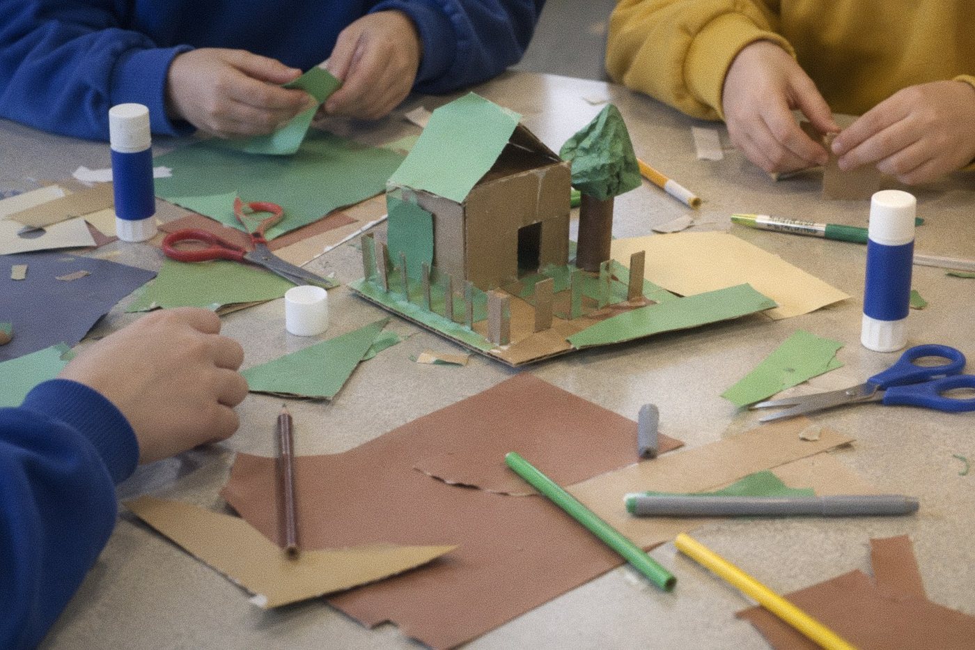 Children's hands cutting and gluing coloured paper around a small cardboard house on a classroom table