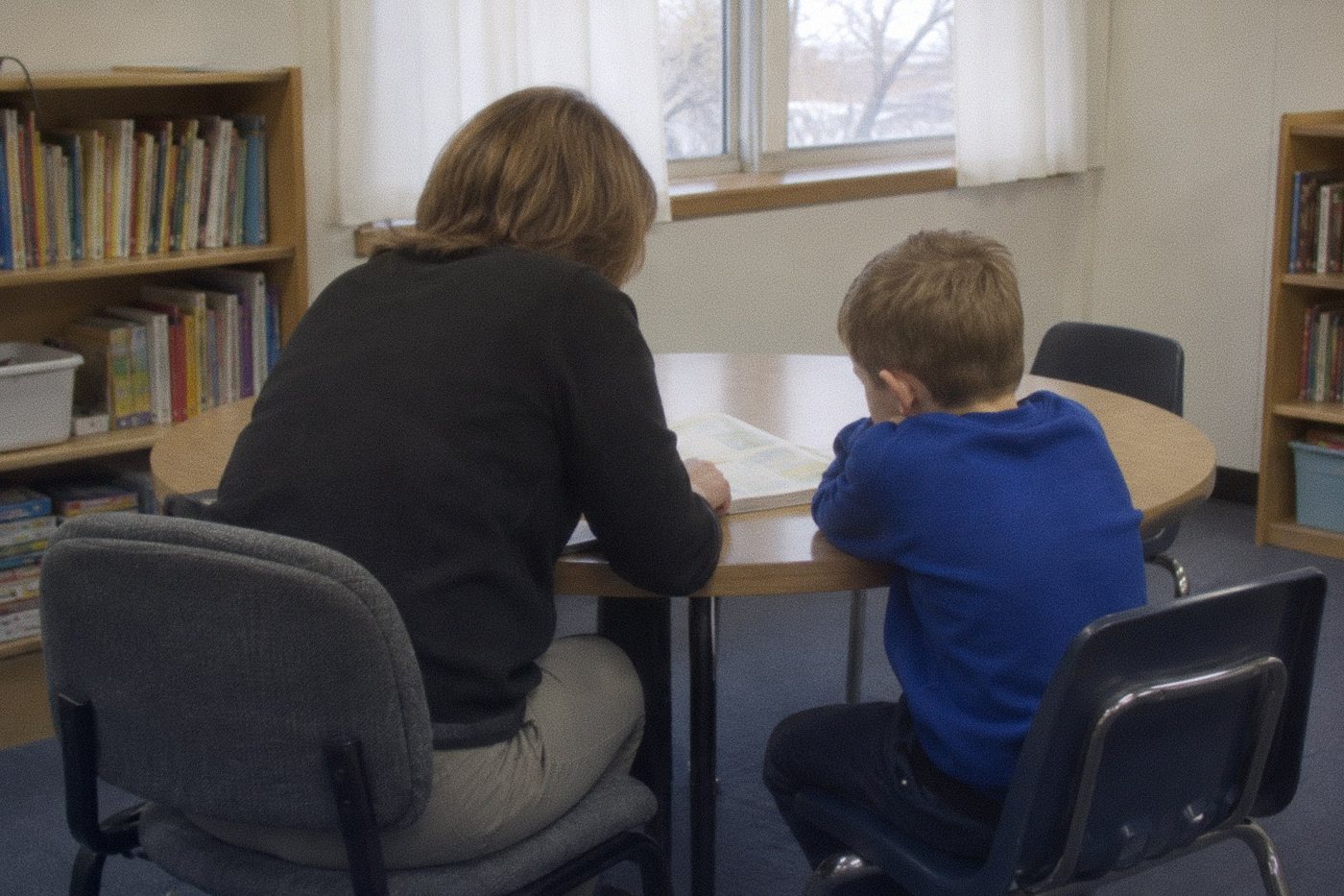 A teacher and a boy in a blue sweatshirt working through a book together at a small round table, seen from behind