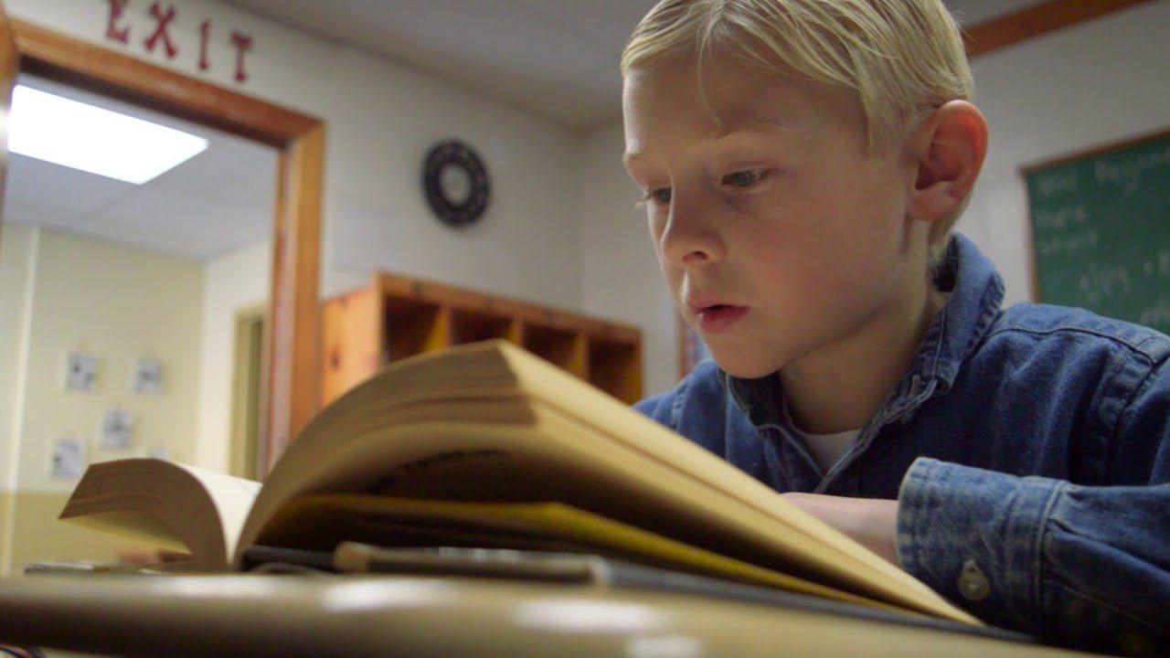 A boy with short blond hair, in a denim shirt, reading a thick open book at a desk in a classroom