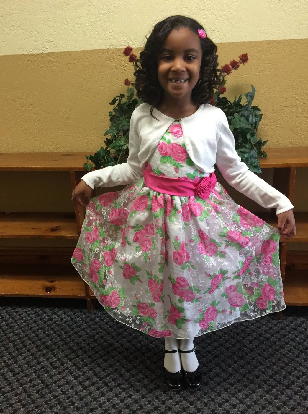 A smiling first-grade girl in a white cardigan and a pink rose dress, in the school hallway