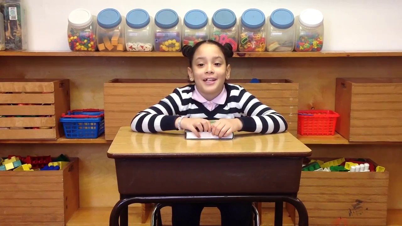 A smiling girl in a striped sweater with her hair in two buns, sitting at a school desk with a small book under her hands, in front of shelves of toys and bins